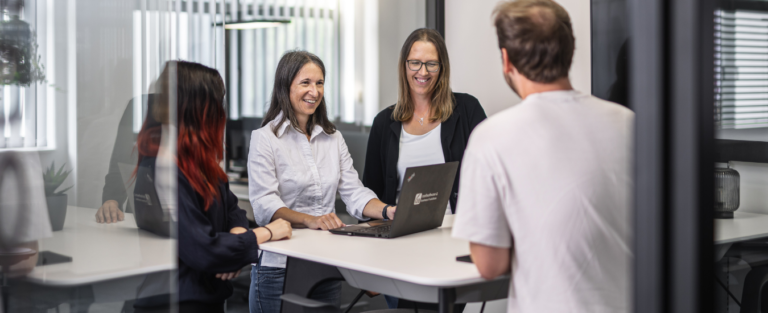 4 Mitarbeiter stehen gut gelaunt bei einem Meetingtisch und unterhalten sich über das Sichern von Daten.